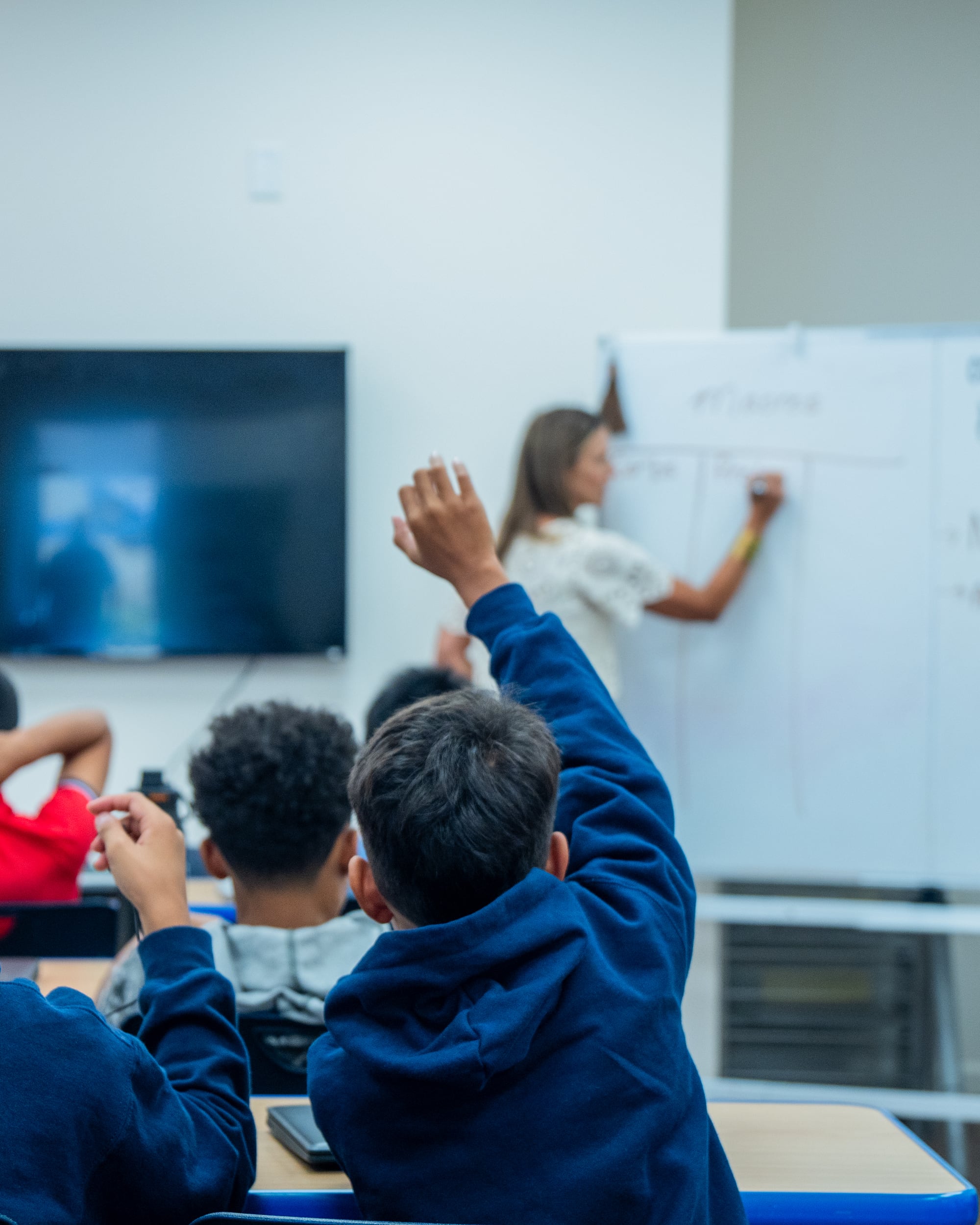 A student in a navy hoodie raises his hand as a teacher writes on a whiteboard in an ISI Academy classroom.