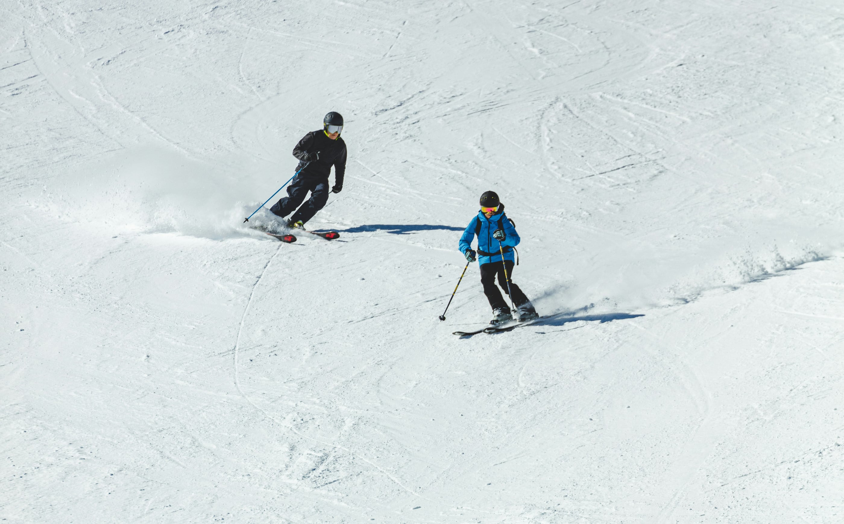 An adult skier leads a young skier down a sunlit slope, the two carving turns side by side.